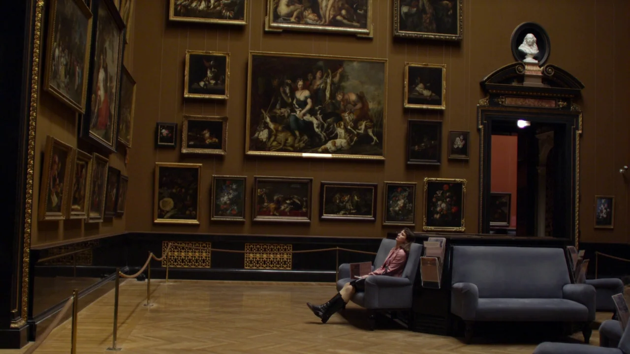 Museum Hours (2012) movie still: wide — A spacious museum gallery features high walls covered in numerous ornate, framed oil paintings. A single…