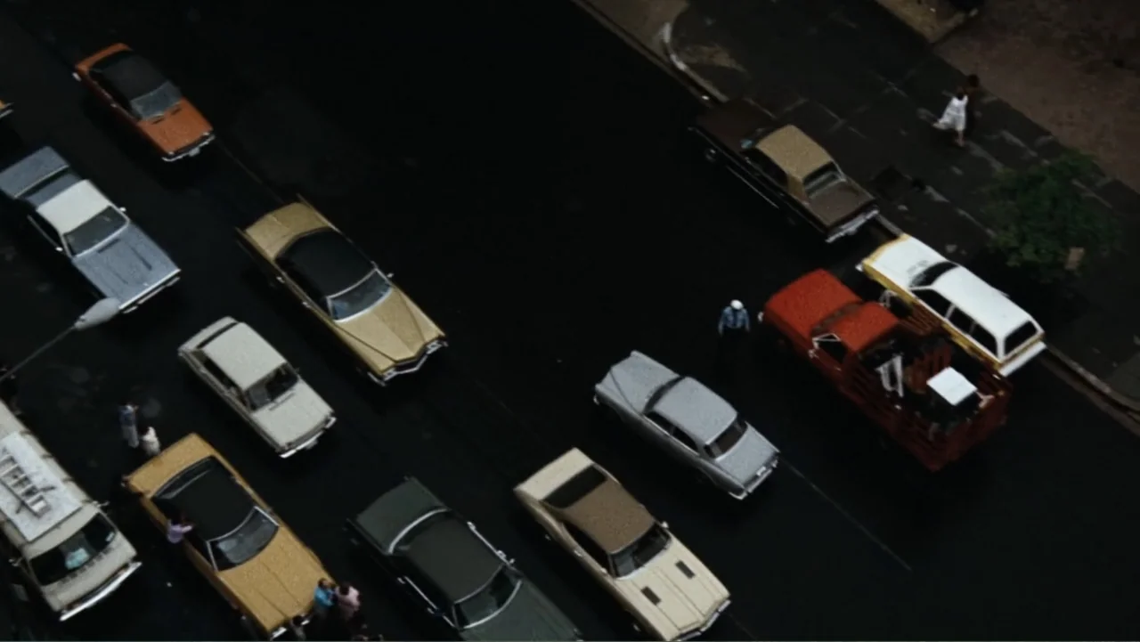 All the President's Men (1976) movie still: establishing — A wet asphalt city street is lined with various mid-1970s model cars. Several vehicles…