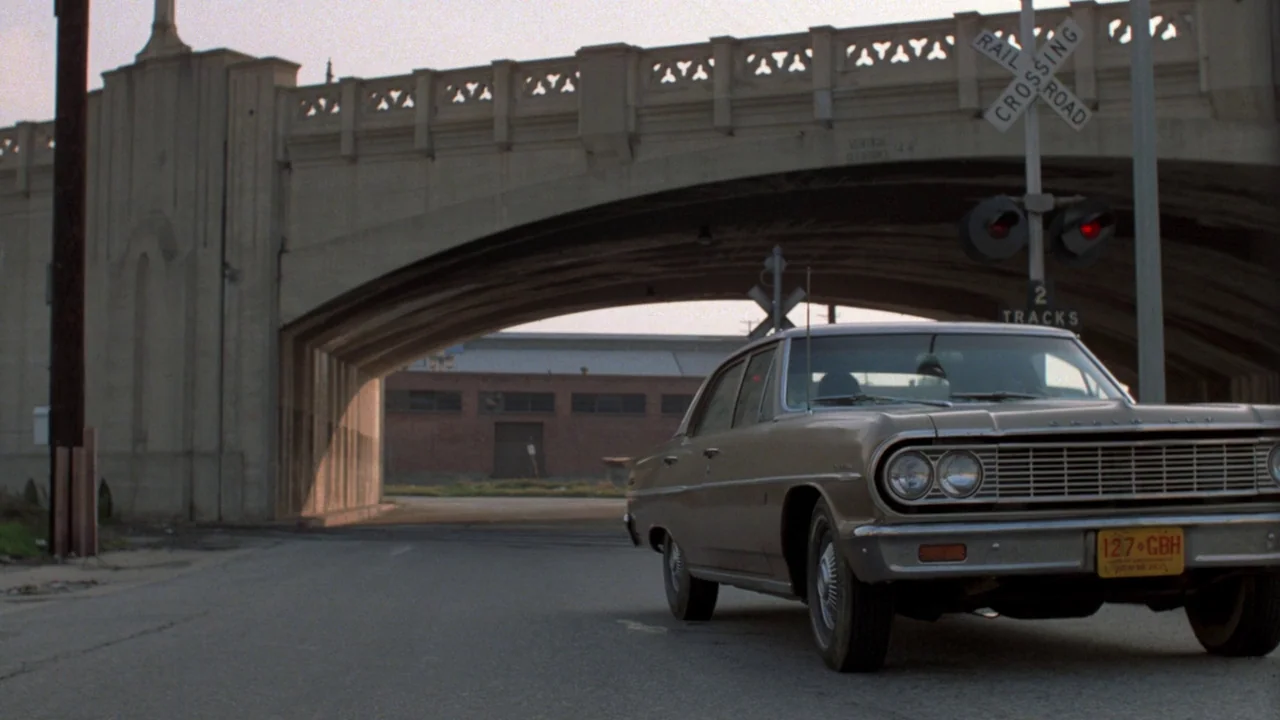 Repo Man (1984) movie still: wide — A concrete railway overpass looms above an asphalt road at an intersection. A tan vintage sedan sits near a rail…