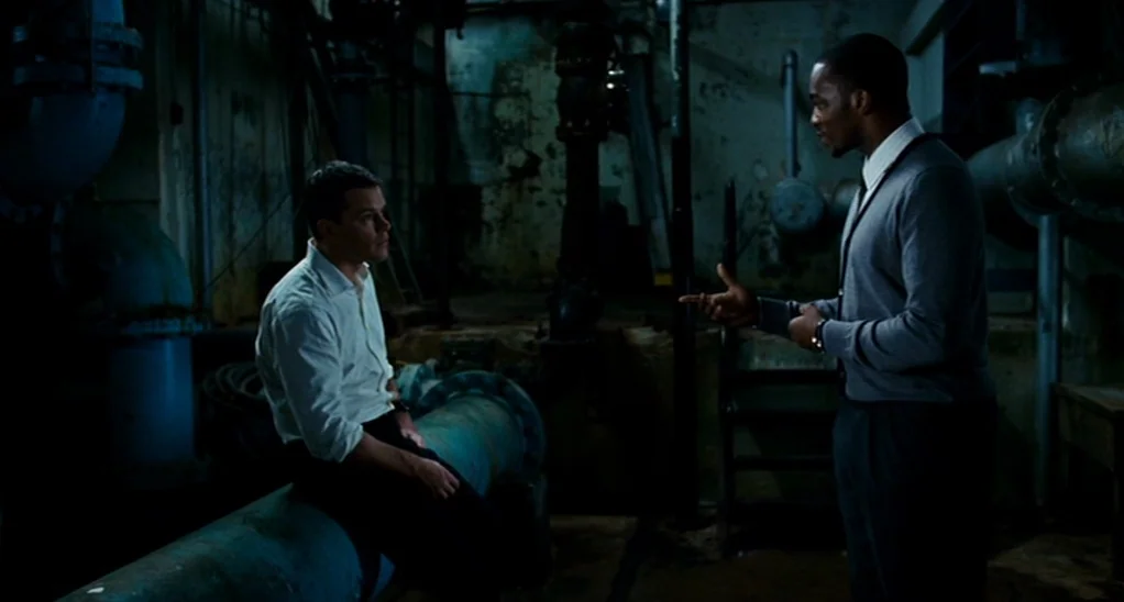 The Adjustment Bureau (2013) movie still: medium — A dimly lit utility room features large metal pipes and dark, industrial concrete walls. Two men…