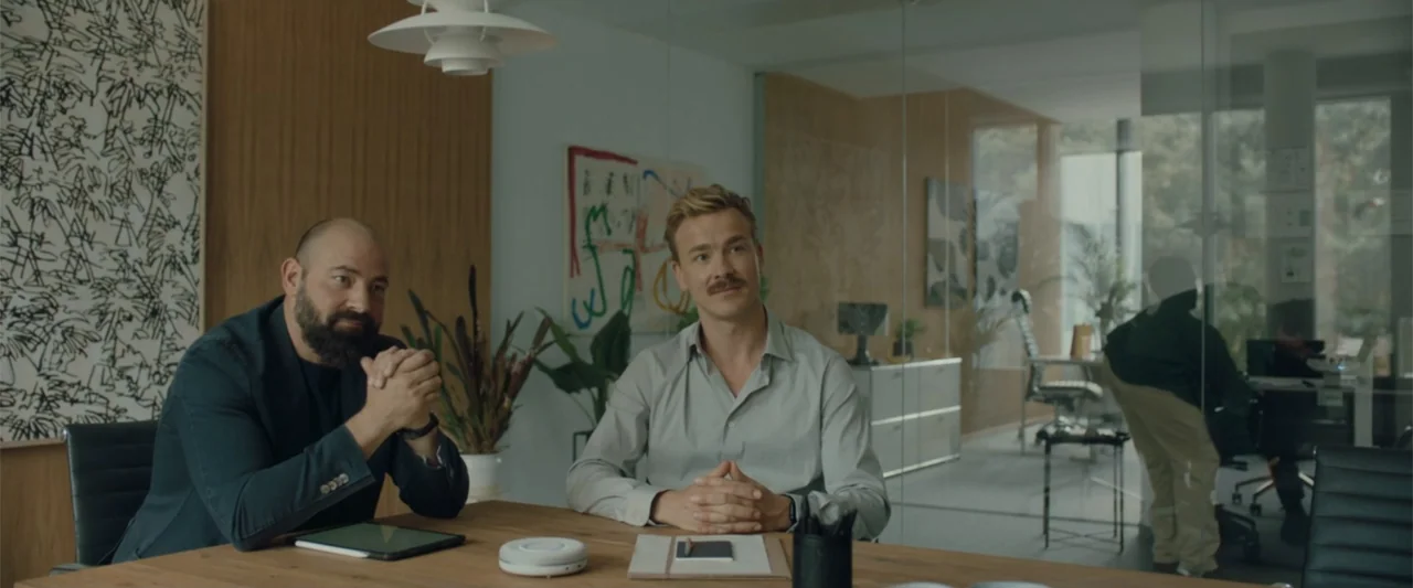 Peacock (2010) movie still: medium — Two men sit at a wooden conference table in a glass-walled office with wood paneling. The subjects sit…