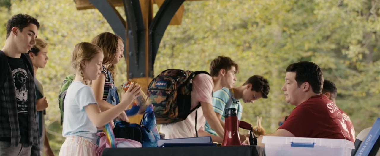 Camp Hideout (2023) movie still: medium — A group of people stand under a wooden pavilion in a wooded park area. A man seated at a table interacts…