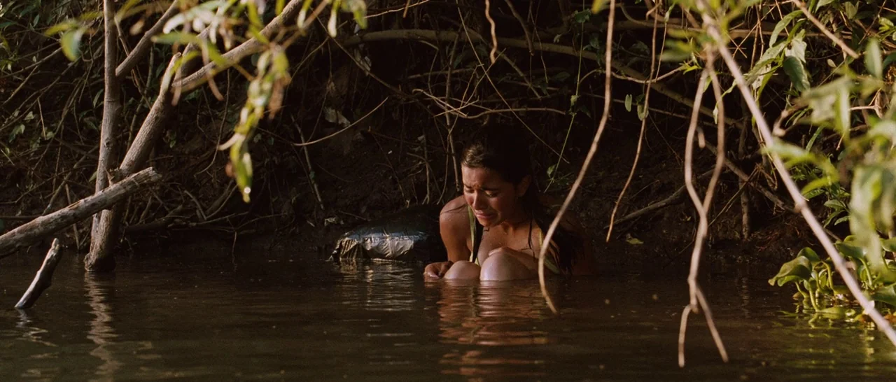 Sin Nombre (2009) movie still: medium — A dense riverbank area with tangled roots, overhanging branches, and murky dark water. A young woman…