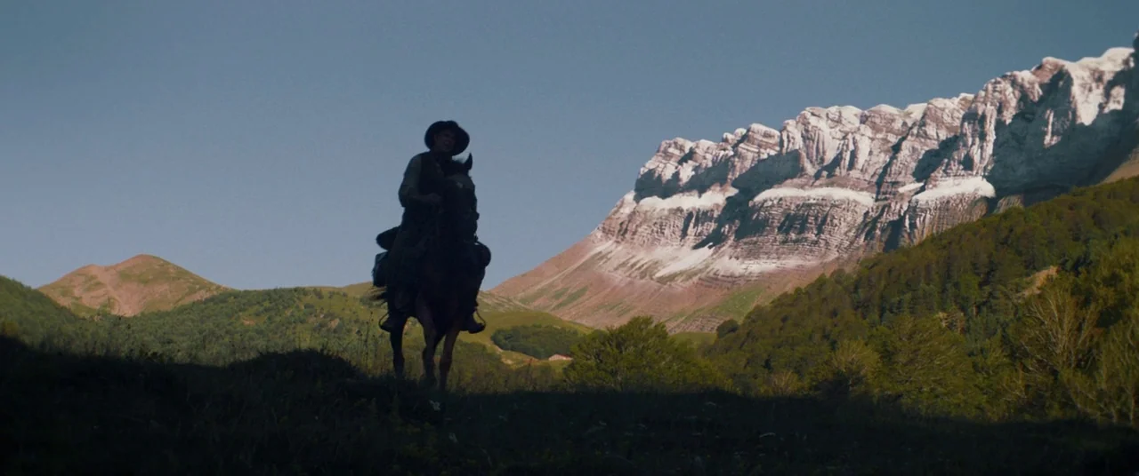 The Sisters Brothers (2018) movie still: wide — A high-altitude rocky mountain range dominates the landscape behind rolling green hills. A lone…