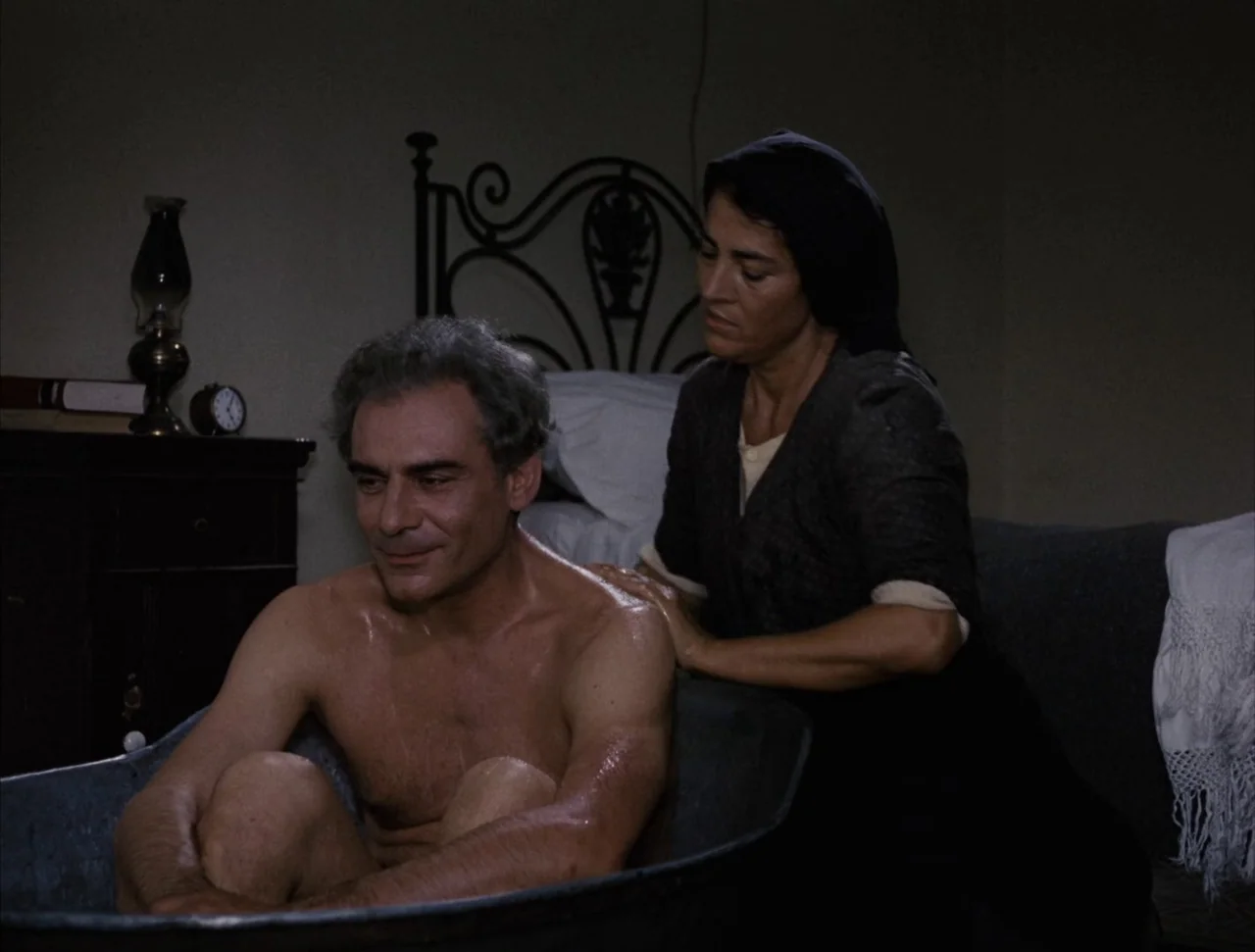 Christ Stopped at Eboli (1979) movie still: medium — A dark, spartan room features a metal wash basin and a simple bed. A woman washes the back of a…