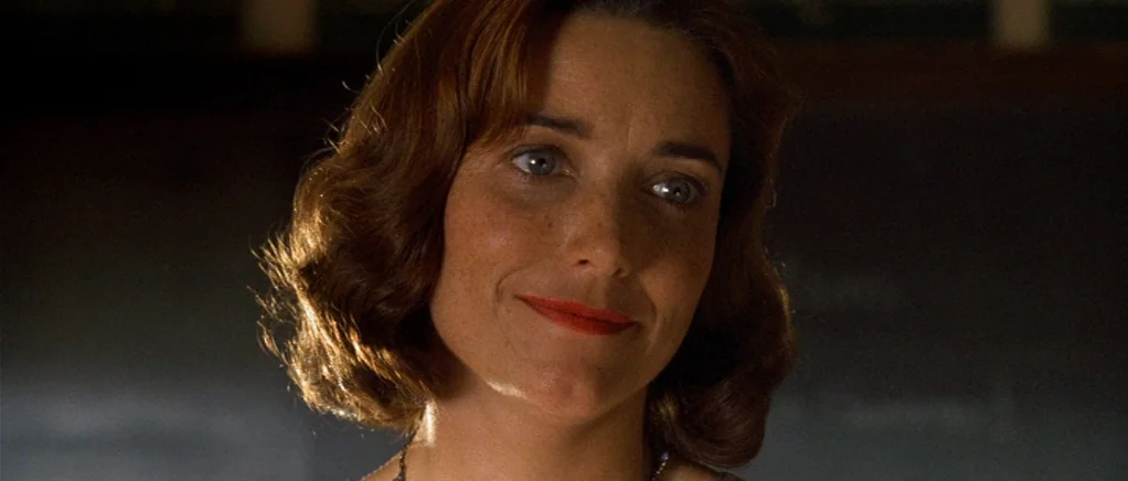 King of the Hill (1993) movie still: closeup — A dimly lit room with a dark board partially visible in the background. A woman with wavy hair looks…