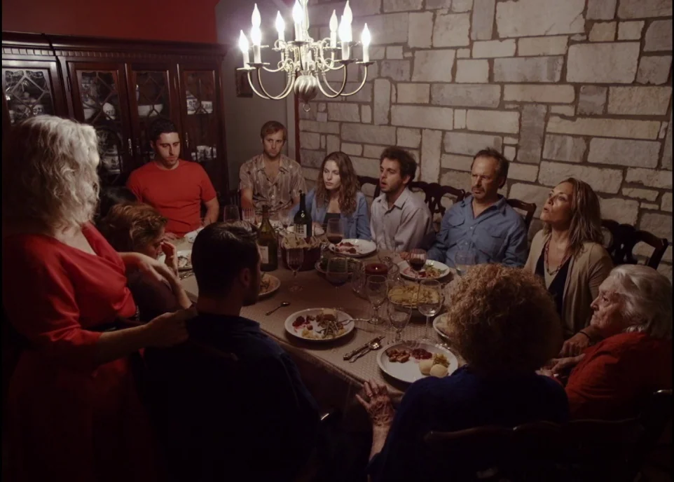Krisha (2015) movie still: wide — A group gathers at a dinner table in a room with stone walls. A chandelier hangs centrally as people eat dinner…