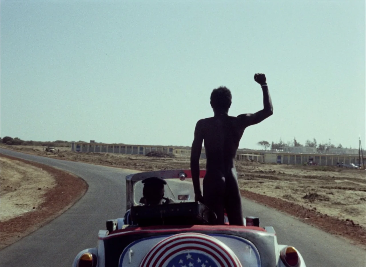 Touki Bouki (1973) movie still: wide — An open asphalt road runs through a dry, arid landscape with distant low buildings. A man stands shirtless in…