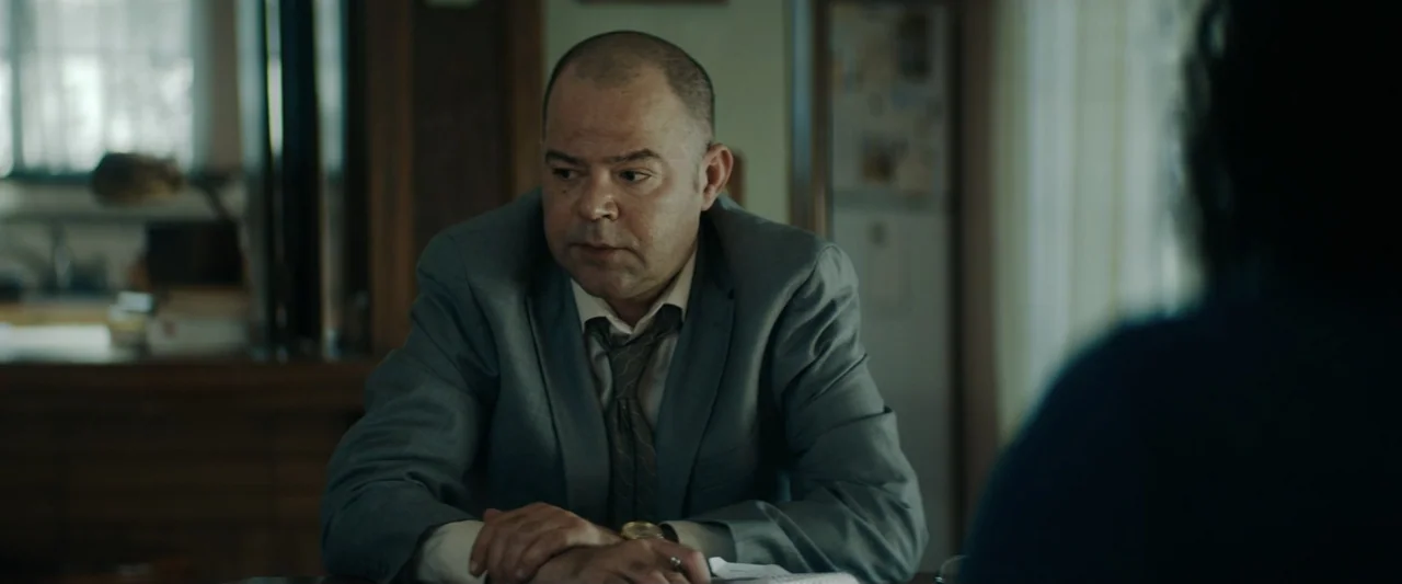 Encounter (2018) movie still: medium — A dimly lit room with plain walls and out-of-focus background furniture. A man in a suit sits at a desk while…