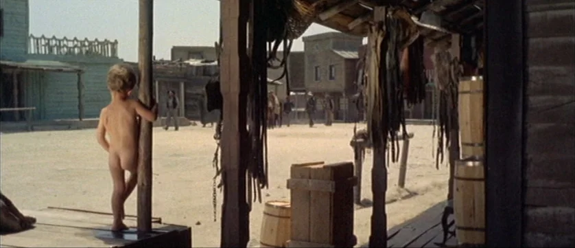 Django Kill (1967) movie still: fullbody — A dusty town square with wooden buildings and a shaded porch in the foreground. A young child stands…