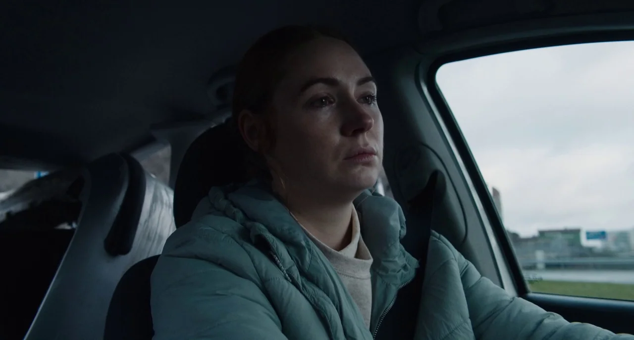Dual (2022) movie still: medium — The interior of a vehicle on an overcast day with blurred outdoor surroundings. A woman in a puffer jacket sits in…