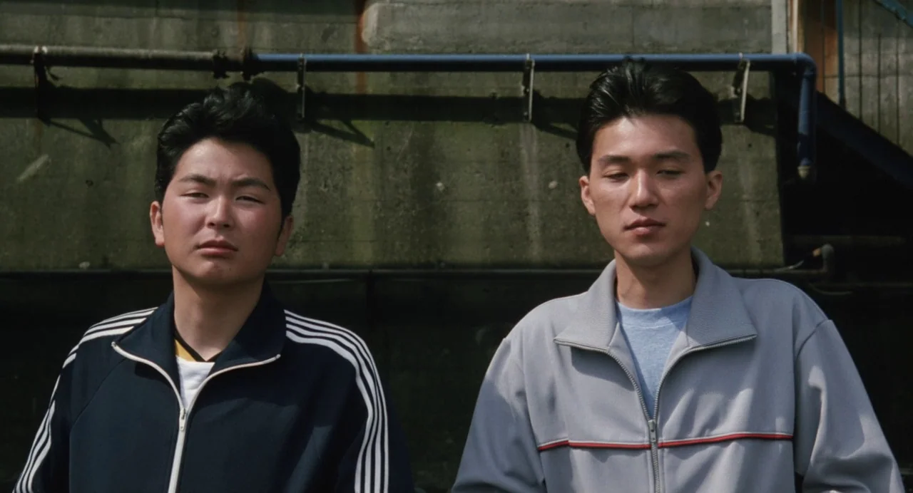 A Scene at the Sea (1991) movie still: medium — Two young men stand in front of a textured, grey concrete wall. They remain still, positioned side…
