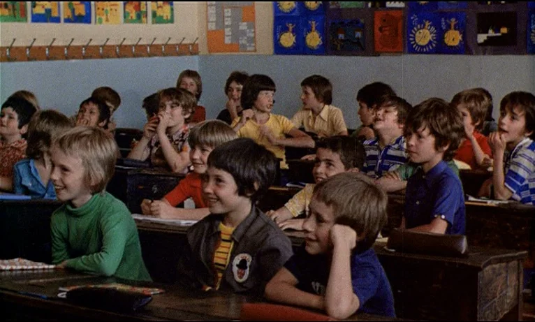 Small Change (1976) movie still: medium — A crowded classroom filled with children sitting in rows of wooden desks. The walls are decorated with…