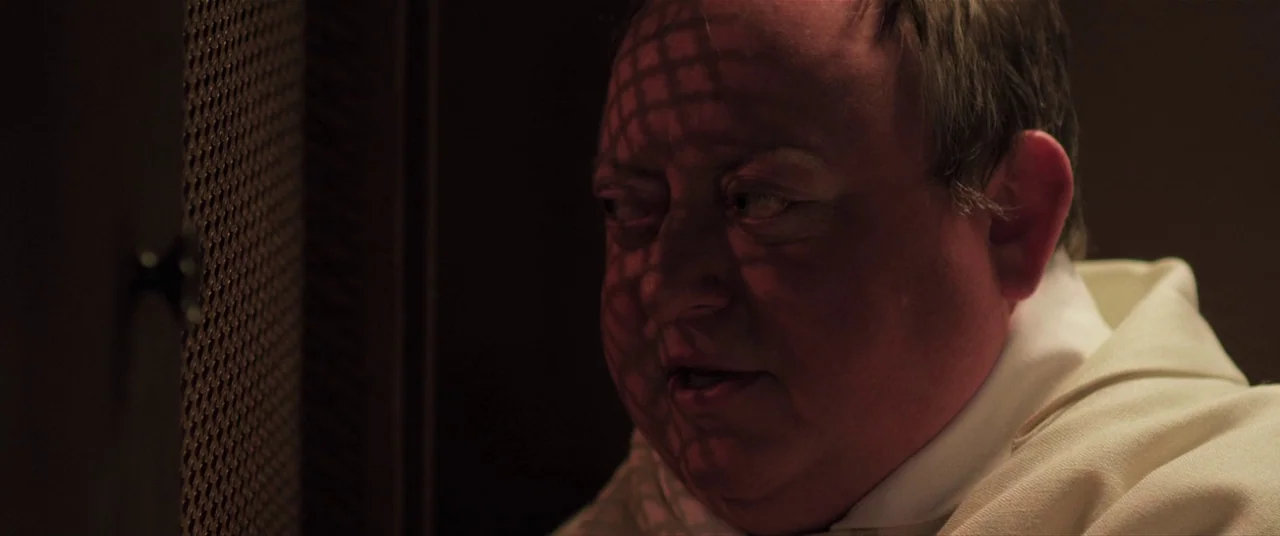 The Editor (2014) movie still: closeup — A dimly lit wooden confessional booth features a metallic lattice screen on the left. An older man in…