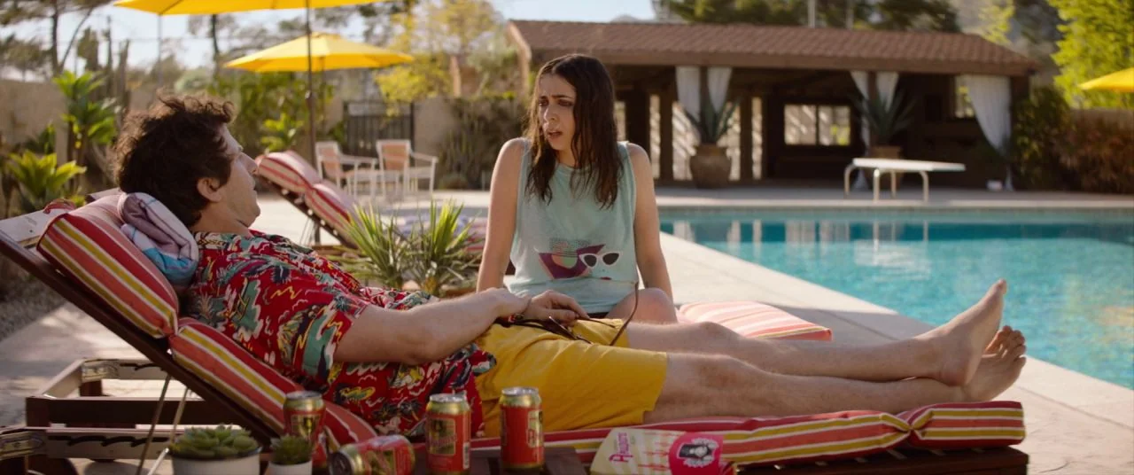 Palm Springs (2020) movie still: medium — A sunny patio area features striped lounge chairs arranged beside a clear blue swimming pool. A man in a…