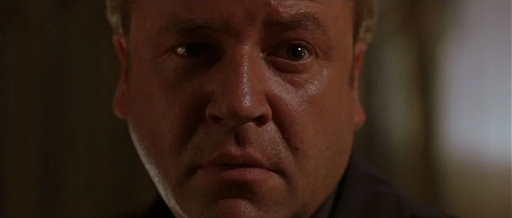 Sexy Beast (2000) movie still: closeup — A dark, nondescript interior space frames a middle-aged man's face. The man stares directly into the lens…