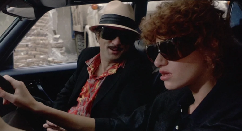The King Of Comedy (1982) movie still: medium — A man and woman sit inside the front seats of a moving car. The driver navigates the road while the…
