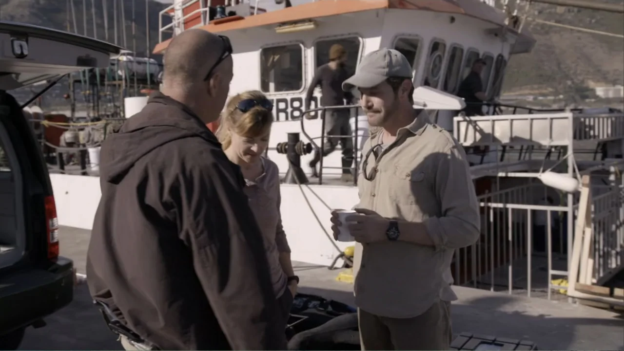 Megalodon: The Monster Shark Lives (2013) movie still: medium — A sunny concrete harbor pier with a large white fishing boat docked in background. A…