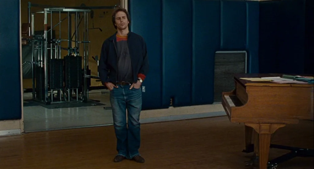 Choke (2008) movie still: fullbody — A multi-purpose room features dark blue padded wall panels and a wooden floor. A man stands in the center near…