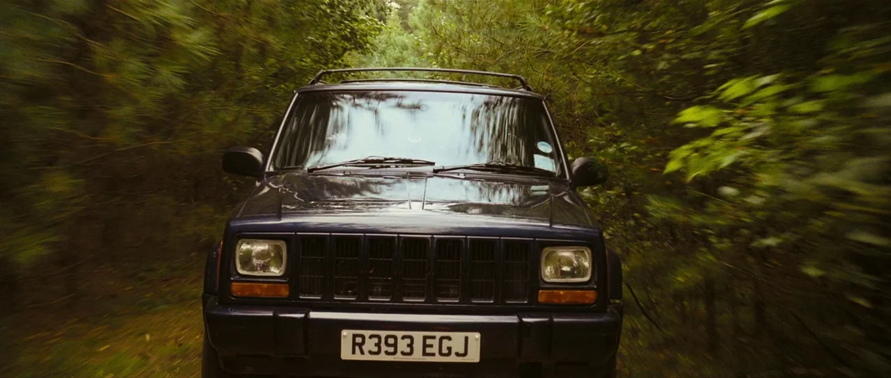 Eden Lake (2008) movie still: medium — A dark blue SUV travels down a narrow, foliage-lined dirt path. The surrounding forest canopy creates a…