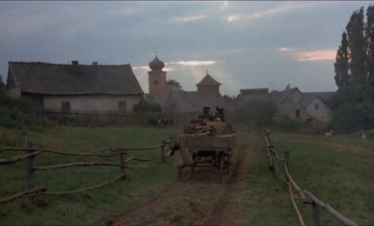 Yentl (1983) movie still: wide — A rural village with small houses and a church steeple sits under a dusk sky. A person steers a horse-drawn cart…