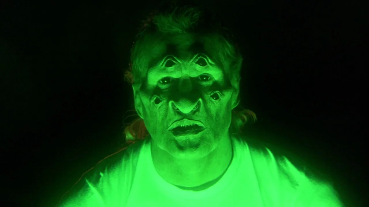 Natural Born Killers (1994) movie still: closeup — A man wearing a multi-eyed creature mask stands in a void of total darkness. The subject stares…