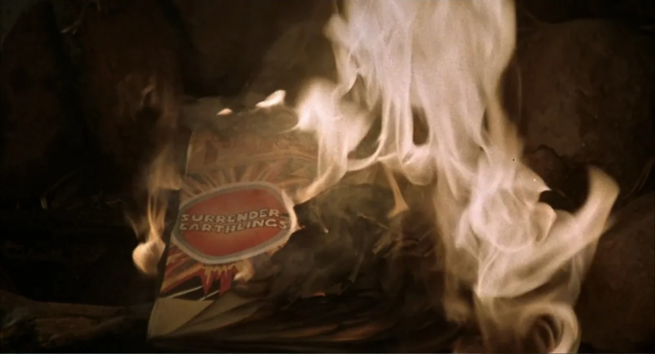 Zone Troopers (1985) movie still: closeup — A close-up view of a paper comic book titled Surrender Earthlings burning in flames. The paper curls and…