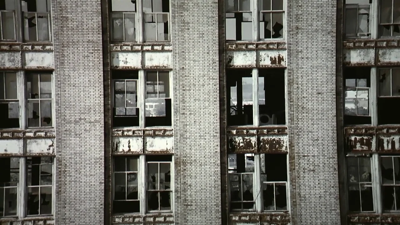Naqoyquatsi (2002) movie still: establishing — The exterior facade of a decaying multi-story industrial building features repeating grid patterns.…