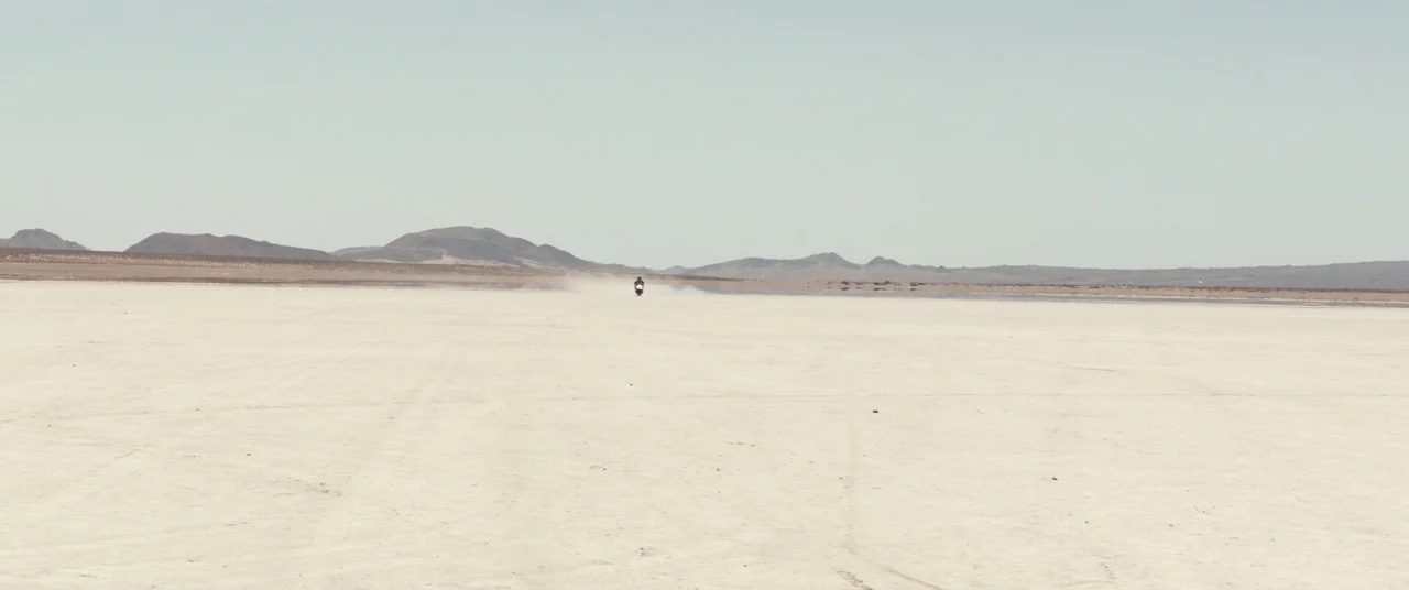 The Bad Batch (2016) movie still: wide — A vast and empty salt flat stretches to a distant mountain range under haze. A lone motorcyclist travels…