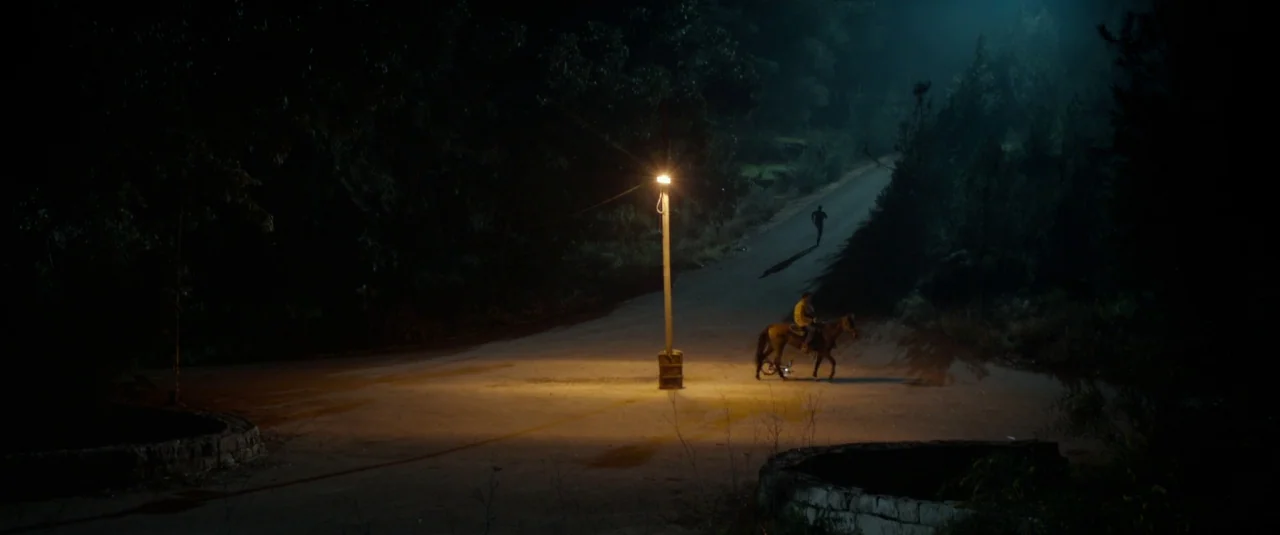 Big Bad Wolves (2013) movie still: wide — A desolate road junction is illuminated at night by a single tall streetlamp. One person rides a horse…