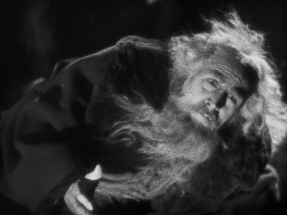 Faust (1926) movie still: closeup — An elderly man lies prone on a dark surface in a dimly lit space. He has a long white beard and messy hair while…