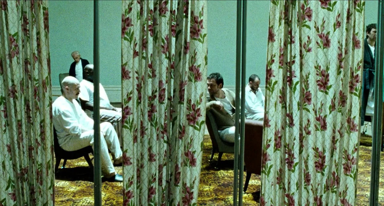 Bronson (2008) movie still: wide — A sterile clinical room features patterned carpet and thin vertical floral room dividers. Several men in simple…