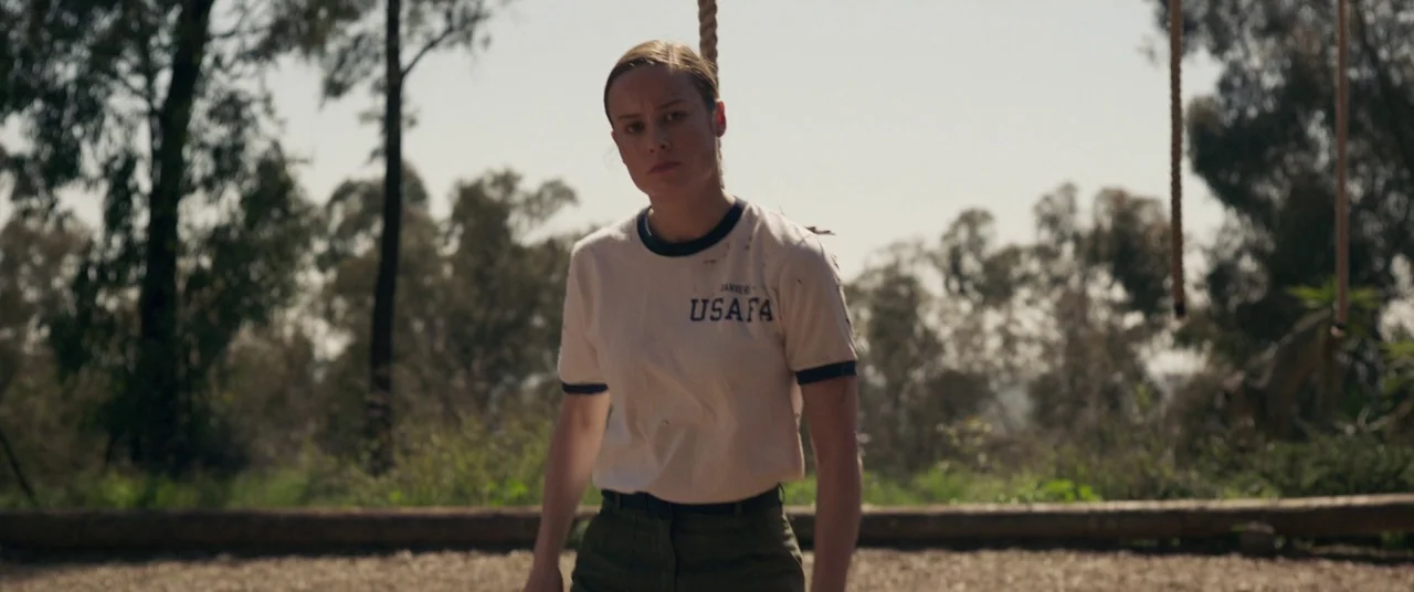 Captain Marvel (2019) movie still: medium — An outdoor obstacle course features dirt ground and vertical ropes under a bright sky. A woman stands in…