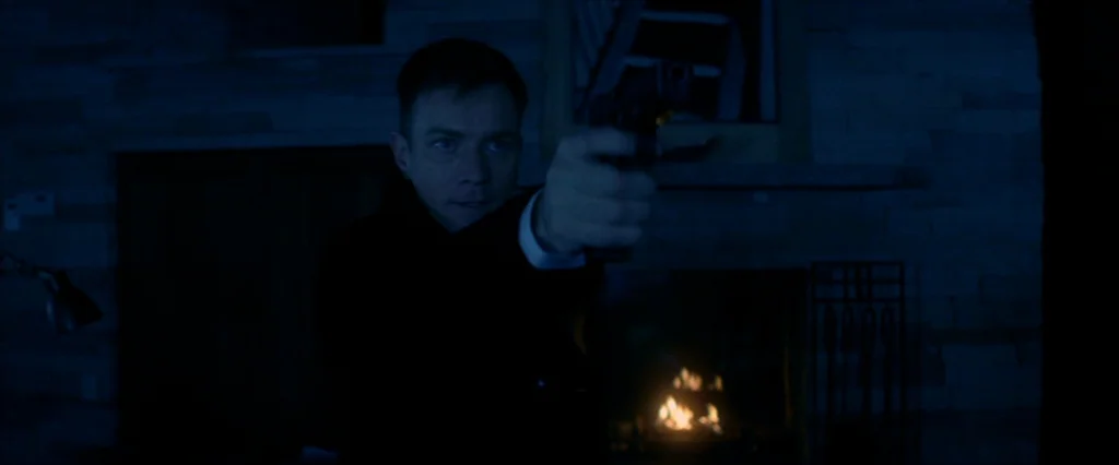 Haywire (2011) movie still: medium — A dark, dimly lit room with a stone fireplace wall as the primary background. A man holds a pistol steady in…