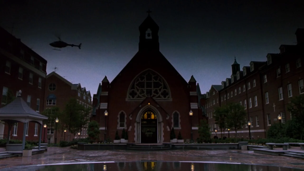 The Exorcist 3 (1990) movie still: establishing — A brick church stands centered in a courtyard at the onset of evening. A helicopter hovers in the…