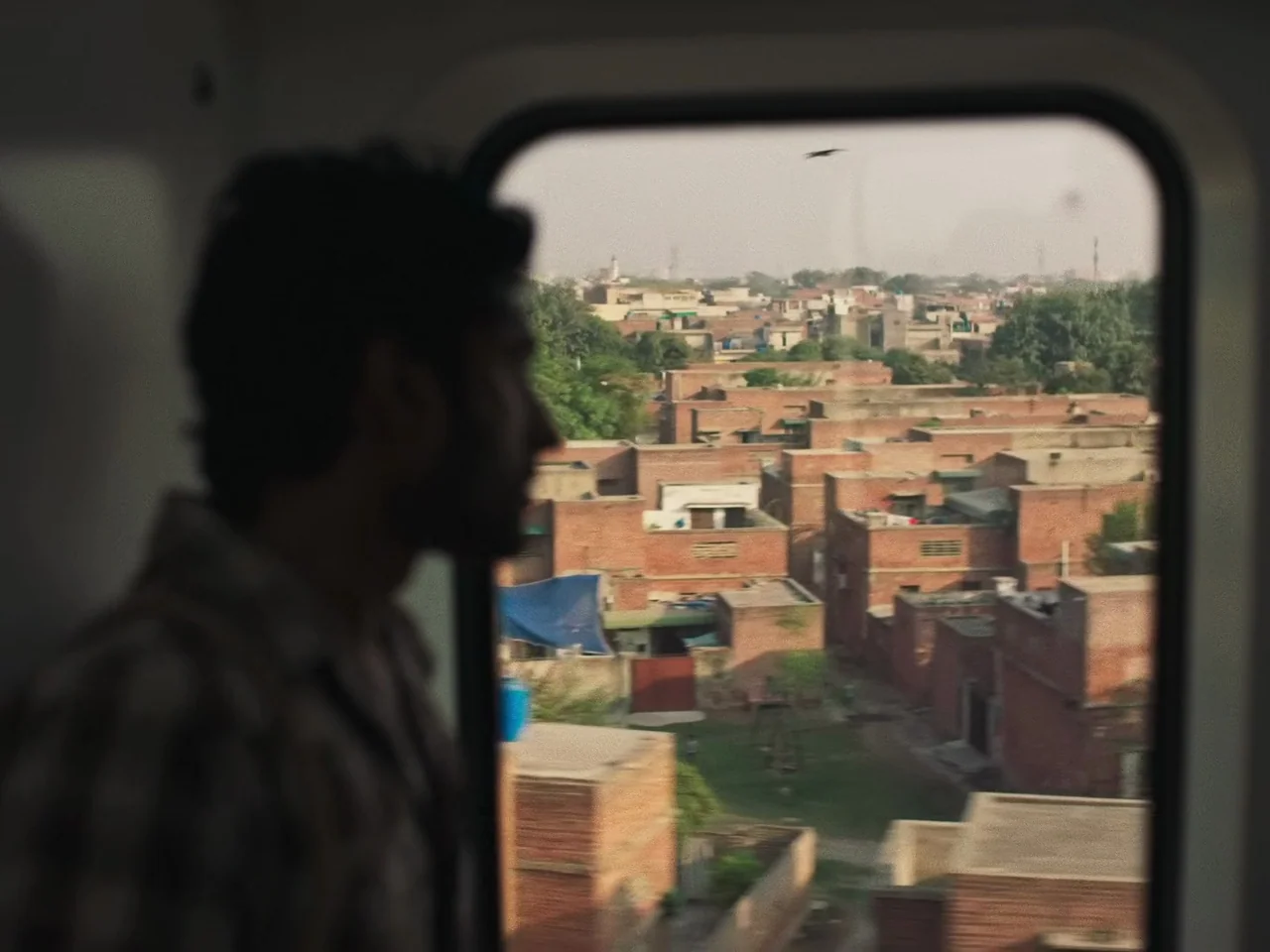 Joyland (2022) movie still: medium — A view from inside a moving train looking out at a densely packed town. Rows of weathered red brick houses and…