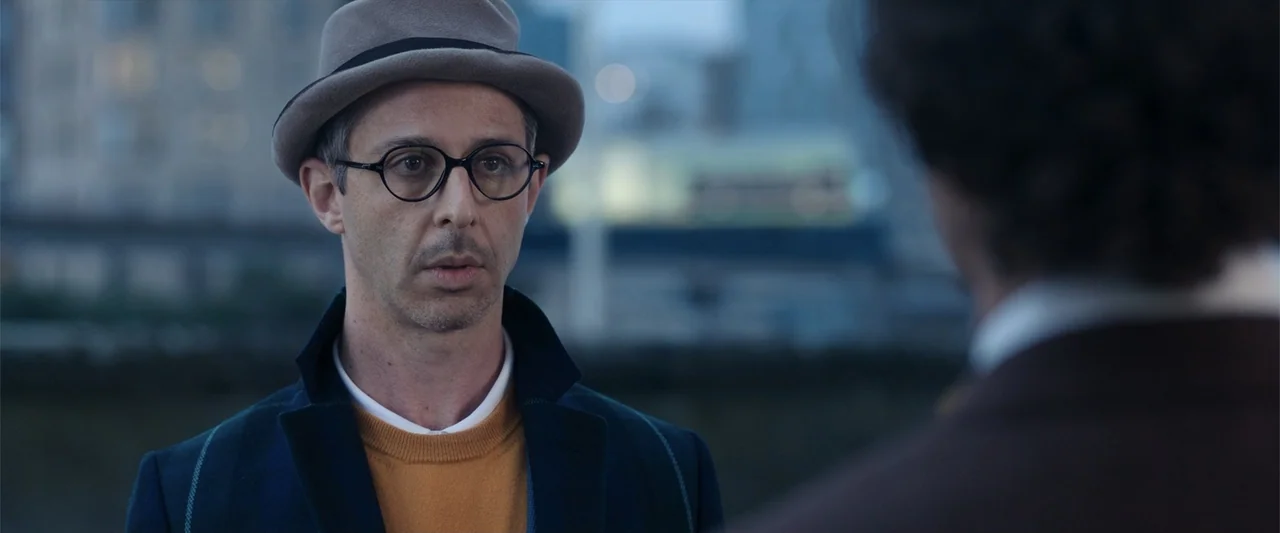 The Gentlemen (2020) movie still: medium — An outdoor riverside area at dusk with a city skyline visible in the distance. A man in a hat looks at an…