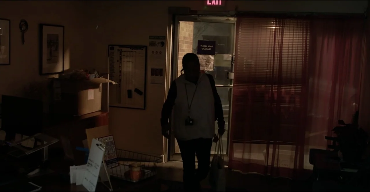 The Devil Is Busy (2024) movie still: medium — A dimly lit office interior contains desk equipment, paper files, and a glass door. A person in a…