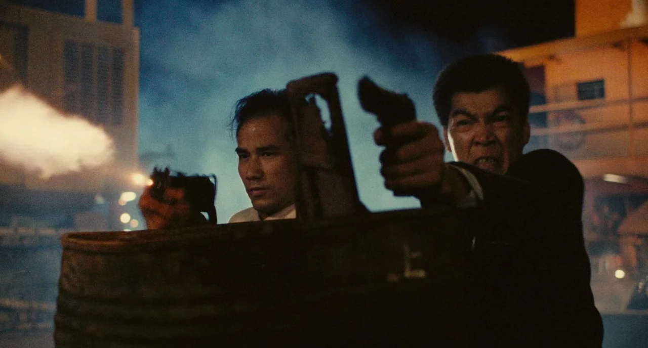 A Better Tomorrow (1986) movie still: medium — Two men fire pistols from behind a weathered metal drum in a nighttime industrial area. Blue smoke…