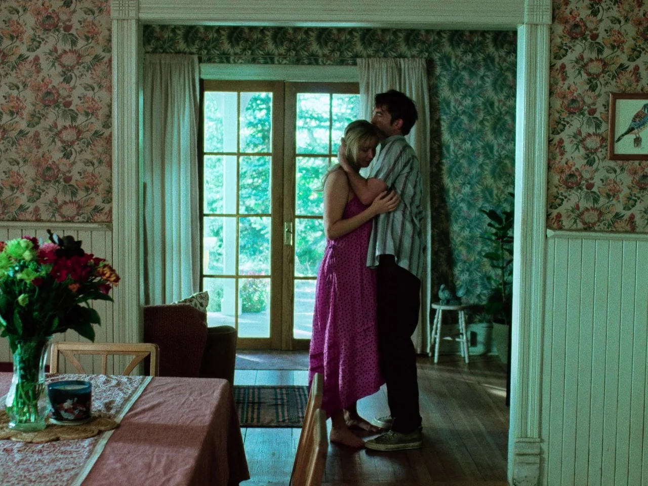 Die My Love (2025) movie still: medium — An interior space features floral wallpaper and a table with a glass flower vase. A man and a woman stand…