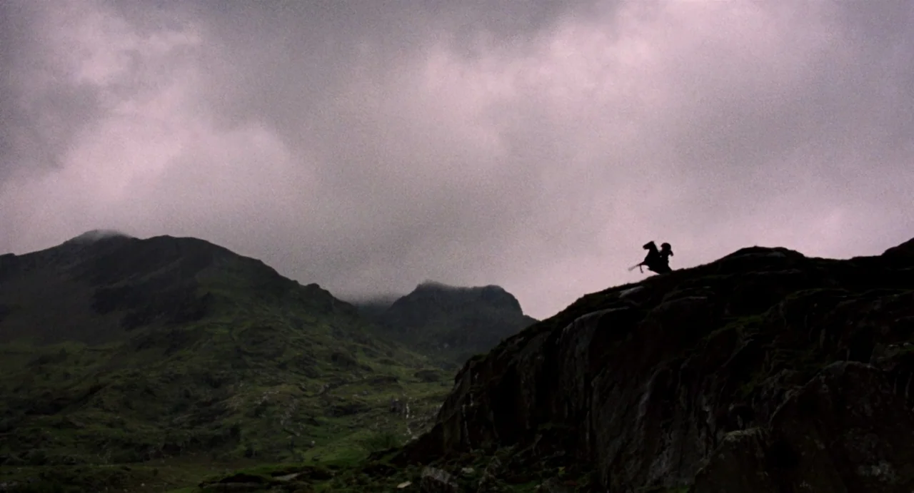 Highlander (1986) movie still: wide — A rugged mountain landscape with rocky terrain and green slopes beneath a cloudy sky. A small, dark silhouette…