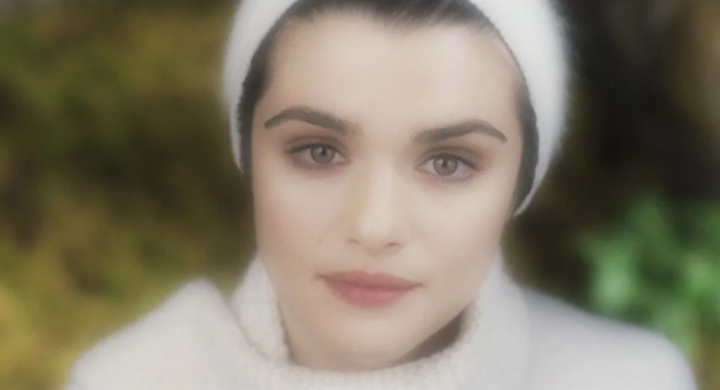The Fountain (2006) movie still: closeup — A woman stands in a blurred outdoor area with green and brown foliage background. She looks directly at…