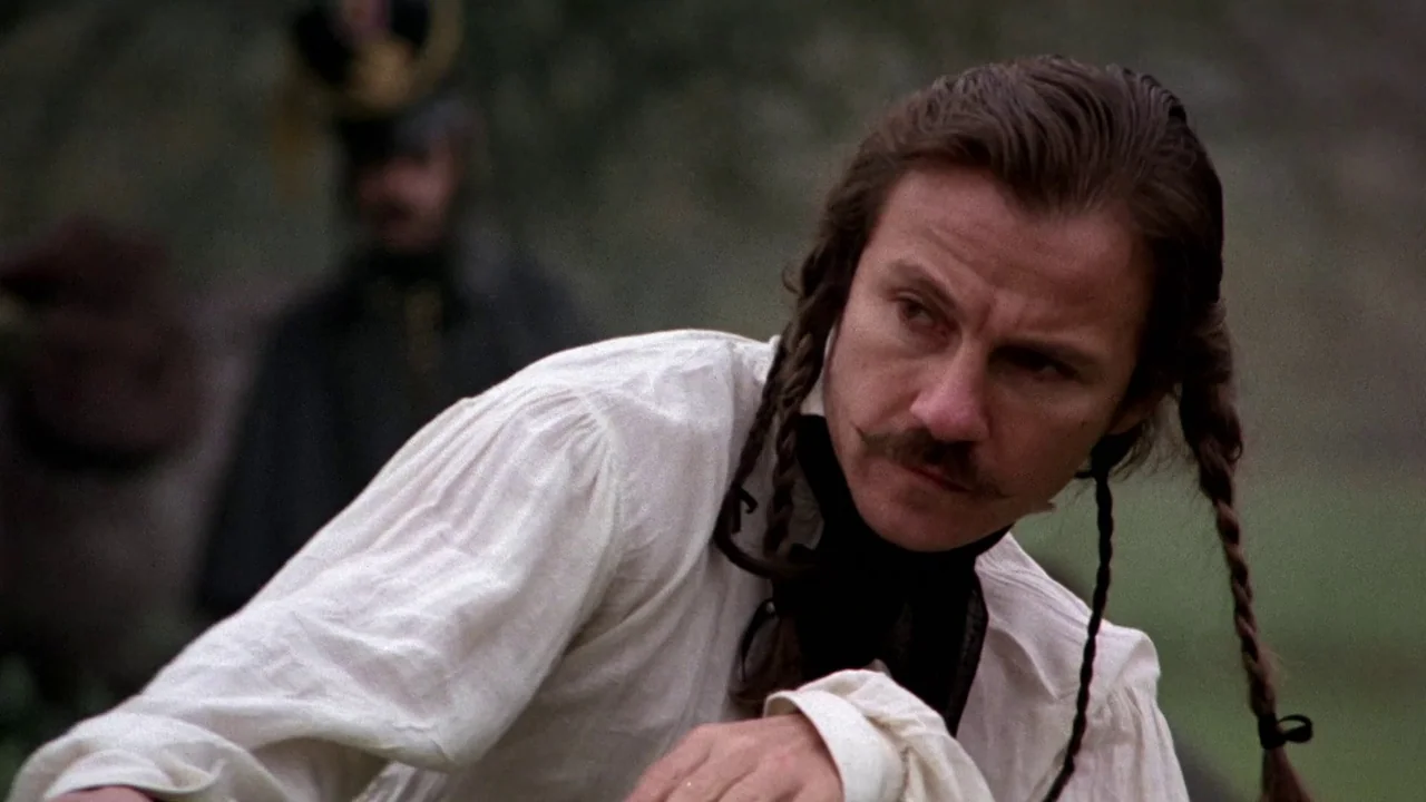The Duellists (1977) movie still: medium — A man with braided hair looks sideways in an outdoor field. In the blurry background, a second figure…