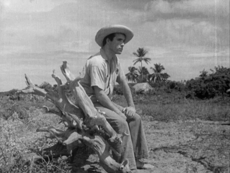 Redes (1936) movie still: fullbody — A man sits on a weathered, exposed tree root in a sparse, dry landscape. Palm trees are visible on the horizon…