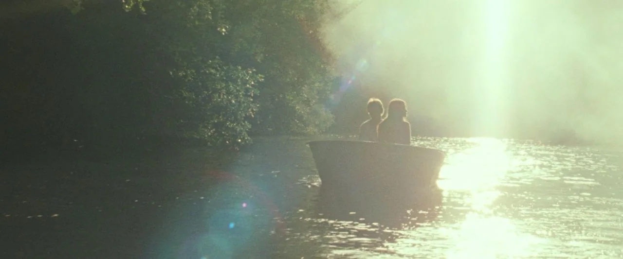 Adoration (2019) movie still: wide — A rowboat carries two people across a calm river near dense green trees. Sunlight reflects off the water…