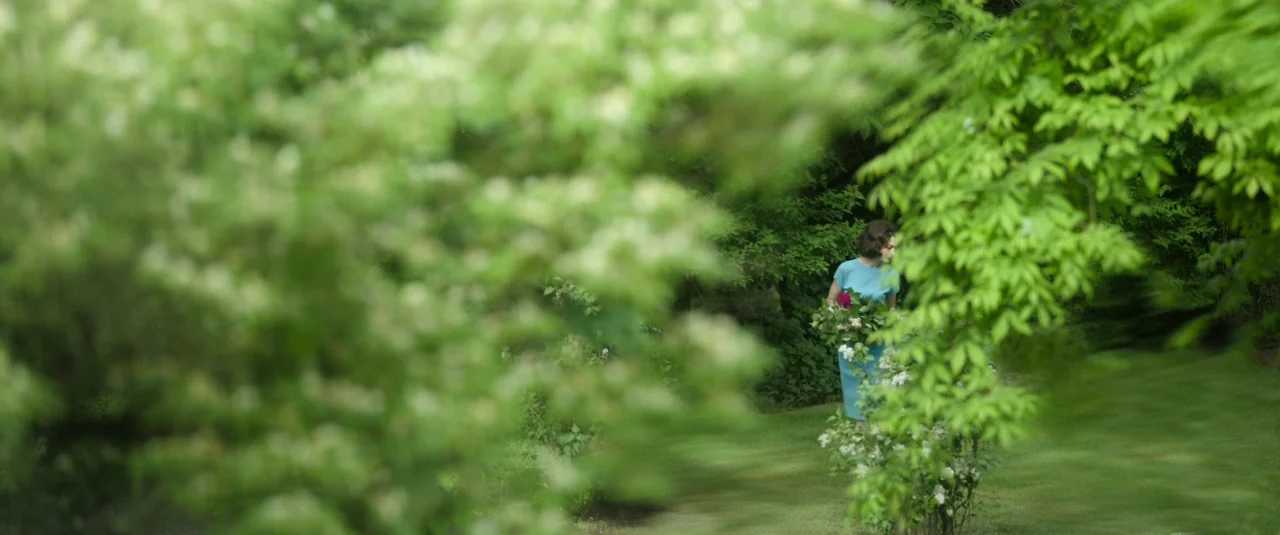 Mother’s Instinct (2024) movie still: wide — A dense garden with soft-focus foliage in the foreground obscuring a walking path. A woman in a blue…