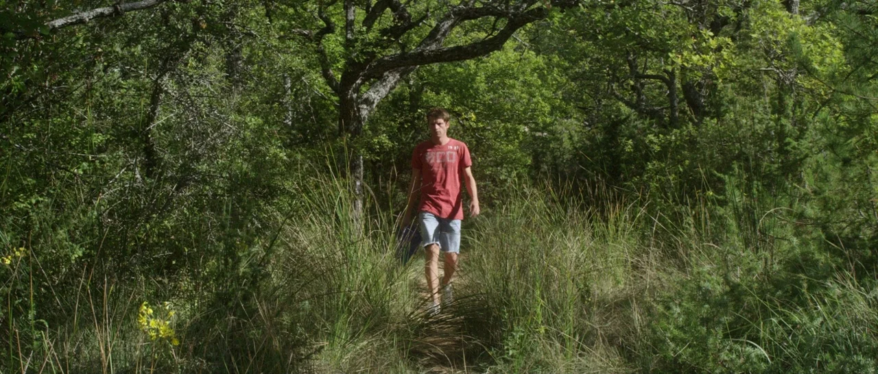 Stranger By The Lake (2013) movie still: fullbody — A man walks along a narrow dirt trail winding through a dense, leafy thicket. High, wild grasses…