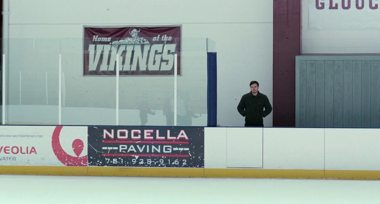 Manchester By The Sea (2016) movie still: wide — An indoor ice hockey rink features a white wall with a banner reading Home of the Vikings. A man…