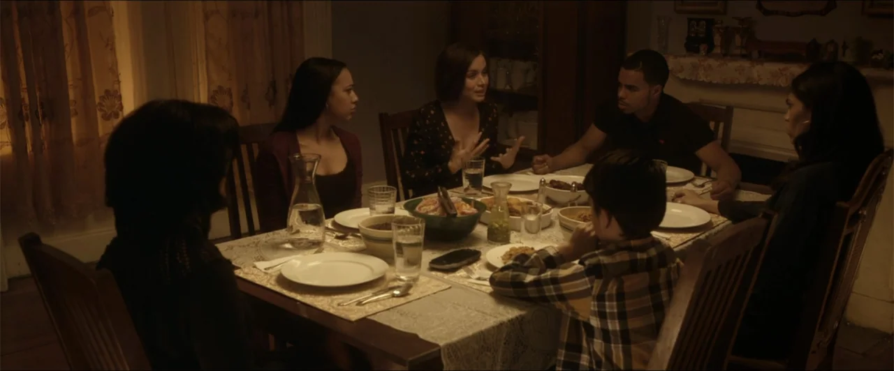 Exposed (2016) movie still: medium — A group of five people gather around a wooden table in a dimly lit home. They are eating a meal while talking…