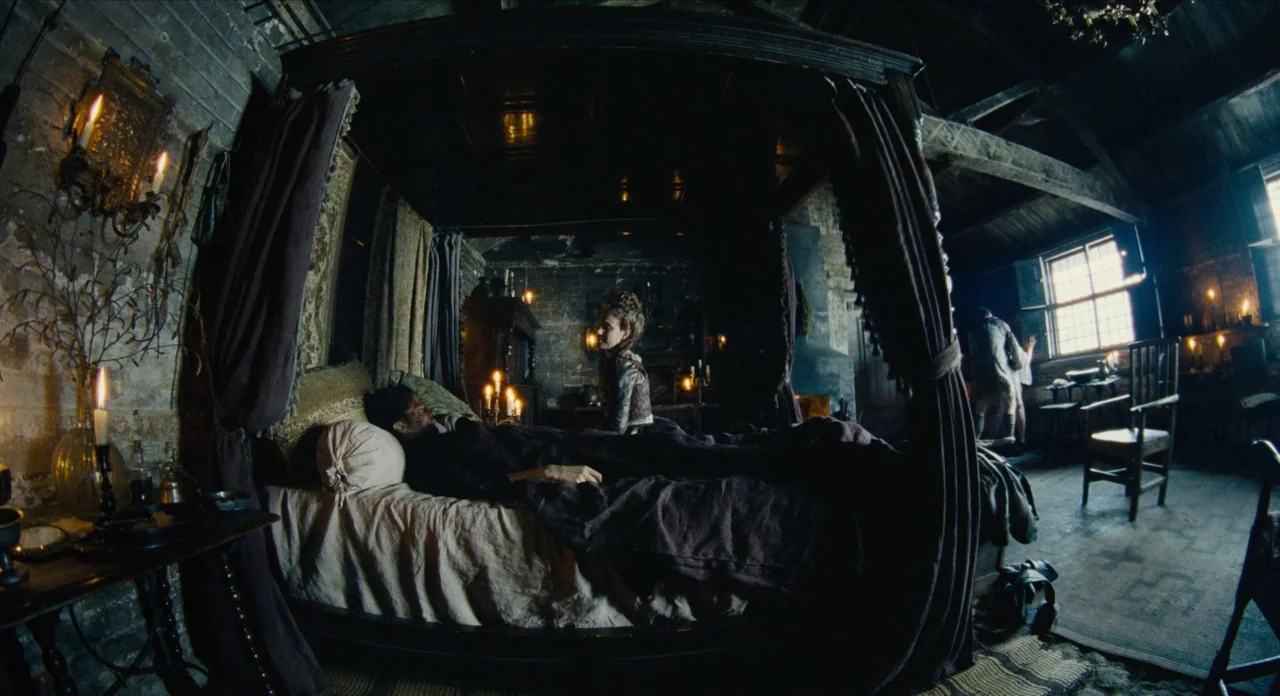 The Favourite (2018) movie still: wide — A dark, wood-paneled room features a large four-poster bed draped in heavy fabric. A woman sits on the edge…